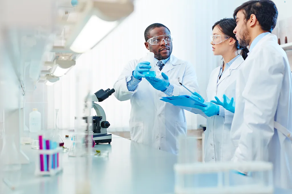 Scientist holding flask discussing chemical reaction with colleagues. Image courtesy of Adobe Stock. 