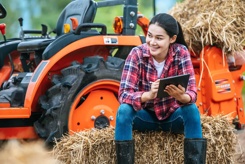 Young female farmer sitting on bale of hay with big tractor machine in farmland, using modern technologies in agriculture management and agribusiness. Courtesy of Adobe Stock