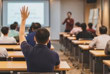 Rear view of audience in raise hand up response for answer the question in a classroom. Image courtesy of Adobe Stock. 